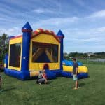 Children and adults are outside on a grassy lawn beside a large, colorful bounce house Long Island on a sunny day, with trees, houses, and a body of water in the background.