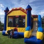 A large colorful inflatable bounce house Long Island with a slide sits on grass. An adult helps a child at the entrance. Trees and a basketball hoop are visible in the background under a blue NY sky with clouds.