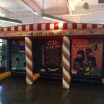 Inflatable carnival games booth with a red and white striped roof, featuring colorful game panels like Star Darts and other toss games, perfect for party rentals Long Island events, set indoors on a shiny floor.