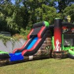 An inflatable zombie-themed bounce house Long Island with a slide sits on grass in a backyard, featuring brick and handprint designs, green and red colors, and a zombie head decoration. Perfect for NY party rentals; trees and a white fence are in the background.