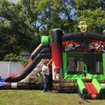 A man stands next to a large zombie-themed bounce house Long Island, featuring a slide on the left. Set up on grass near white fences and tall green trees, this striking inflatable is perfect for NY party rentals Long Island.