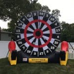 An oversized inflatable dartboard with three velcro soccer balls stands on grass in a fenced NY backyard, perfect for party rentals Long Island, with trees and a house completing the fun scene.