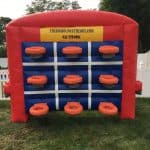 An inflatable tic-tac-toe game with blue and red colors stands on grass in a fenced backyard. Perfect for party rentals Long Island, NY, it features orange rings on the grid and a yellow sign with contact information at the top.