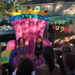 Two girls sit on a large, inflatable pink princess throne—available from party rentals Long Island, NY—at an outdoor birthday party. Balloons and a “Happy Birthday” banner add to the festive scene surrounded by children.