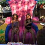 Two young girls sit on a large, colorful inflatable throne outdoors at a birthday party in NY. A birthday cake with candles and decorations sits on the table in front of them, showing off the fun made possible by party rentals Long Island.