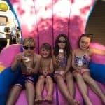 Four children in swimsuits sit together on a large, colorful inflatable chair from a party rentals Long Island company, enjoying ice cream cones on a sunny day. They appear happy and relaxed, with a camper and balloons in the background.