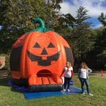 Two women stand in front of a large inflatable pumpkin bounce house Long Island, featuring a jack-o’-lantern face, set up outdoors on the grass under a partly cloudy NY sky—perfect for party rentals Long Island events.