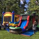 Two colorful inflatable bounce houses, including one with a slide, are set up on grass in a park. Several children and adults stand nearby under tall trees and blue sky—perfect for party rentals Long Island or NY gatherings.