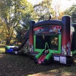 A large inflatable bounce house Long Island with a zombie theme, featuring bright colors, cartoon zombies, a slide on the left, and surrounded by trees in a grassy outdoor area—perfect for NY party rentals.