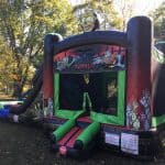 A large inflatable zombie-themed bounce house, perfect for party rentals Long Island, features cartoon zombies and the word "ZOMBIES" above the entrance. Set up outdoors on grass in NY, it's surrounded by trees in daylight.