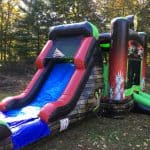 A colorful inflatable bounce house Long Island with a slide is set up outdoors on grass, surrounded by trees. The bounce house features a stone wall pattern, bright red, green, and blue sections, and spooky handprints on one side.