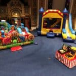 Colorful inflatable bounce houses and slides—ideal for party rentals Long Island—are set up inside a large, ornate hall with decorative walls and blue carpet. A small carnival game booth is in the foreground; a table sits in the background.