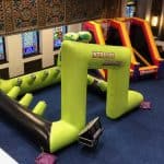 An indoor setup featuring large, colorful bounce house Long Island obstacle courses on a blue carpet, with stained glass windows and ornate wall designs in the background. The main inflatable reads “Interactive Challenge”—perfect for party rentals Long Island, NY.