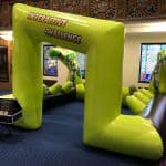Bright green inflatable game setup labeled "Interactive Challenge" with a cartoon character, digital score display on a small table—all part of our bounce house Long Island party rentals—situated on blue carpet inside a room with stained glass windows.