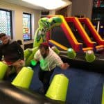Two men play an interactive game with glowing buttons on a bounce house Long Island, NY obstacle course, surrounded by stained glass windows and colorful decorations.