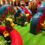 Colorful inflatable obstacle course with cartoon designs, including frogs and butterflies, set up indoors with people and tables in the background. Perfect for party rentals Long Island, its bright shapes create a playful scene kids will love.