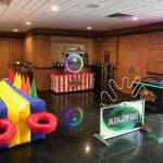 A game room with colorful inflatable ring toss, a MegaWire game, a Touchdown Strike game, and a TAITO arcade machine on a red-and-white striped table—perfect for party rentals Long Island, NY—all set on a shiny black floor near brick walls and doors.