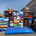 A large inflatable pirate ship bounce house Long Island is set up outdoors near a building, featuring pirate figures, skull flags, and a colorful treasure chest. A person stands beside the red and blue striped entrance—perfect for party rentals Long Island, NY.