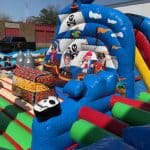 Colorful inflatable pirate-themed bounce house Long Island with cartoon pirates, treasure chests, and skull decorations. Bright blue, red, and green sections pop against the backdrop of a black pickup truck and nearby buildings.