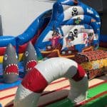 A colorful inflatable pirate-themed play area in NY with cartoon sharks, a treasure chest, a striped arch, and a pirate ship backdrop—perfect for bounce house Long Island party rentals!.
