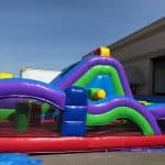 A colorful inflatable obstacle course with arches and climbing areas, featuring red, blue, green, purple, and yellow sections, set up outdoors near a large building—perfect for party rentals Long Island events.