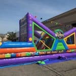 Colorful inflatable obstacle course with vibrant geometric patterns and a “Come One Come All” sign, set up outdoors on a paved surface near a building. Perfect for party rentals Long Island, with small flags hanging across the top.