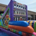Colorful inflatable obstacle course with vibrant patterns and a facade resembling a painted building, available from party rentals Long Island, set up outdoors near a beige building with metal awnings and a clear sky in the background.