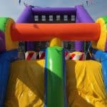 Colorful inflatable obstacle course outdoors with bright yellow, green, orange, blue, and purple sections—perfect for party rentals Long Island, NY. A building and blue sky are visible in the background along with fun red-and-white barriers.