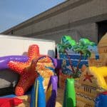 Colorful inflatable structures with ocean and beach themes, including a starfish, fish, palm trees, and a sandcastle—perfect for NY events. Set up outdoors near a white trailer and building wall by party rentals Long Island.
