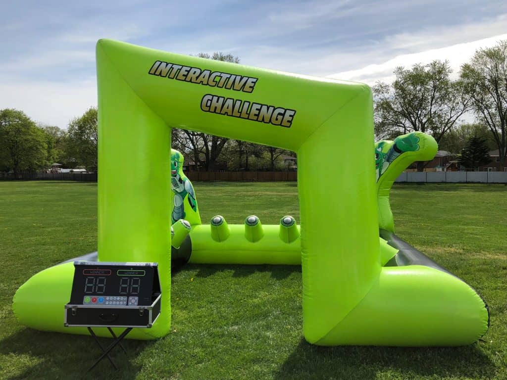Bright green inflatable game structure labeled "Interactive Challenge" stands on grass, perfect for party rentals Long Island. Four targets are inside, with a scoring console on a table to the left. Trees and a fence are visible in the background.