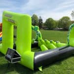 A large, inflatable green and black bounce house Long Island game setup with cartoon hero graphics on each side, placed outdoors on grass. A digital timer is attached to the front left corner. Trees and another inflatable are in the background.