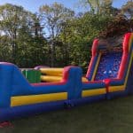 A large, colorful inflatable obstacle course with climbing, sliding, and crawling sections sits on green grass, surrounded by trees on a sunny day—perfect for bounce house Long Island parties or party rentals in NY.