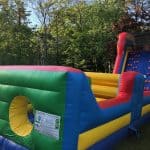A colorful inflatable obstacle course with climbing holds and circular entry holes is set up outdoors on grass, surrounded by green trees and sunlight—perfect for party rentals Long Island and NY celebrations.
