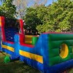 A colorful inflatable obstacle course with climbing steps and tunnels, available through party rentals Long Island, is set up on a grassy lawn surrounded by green trees and a sunny, clear sky.