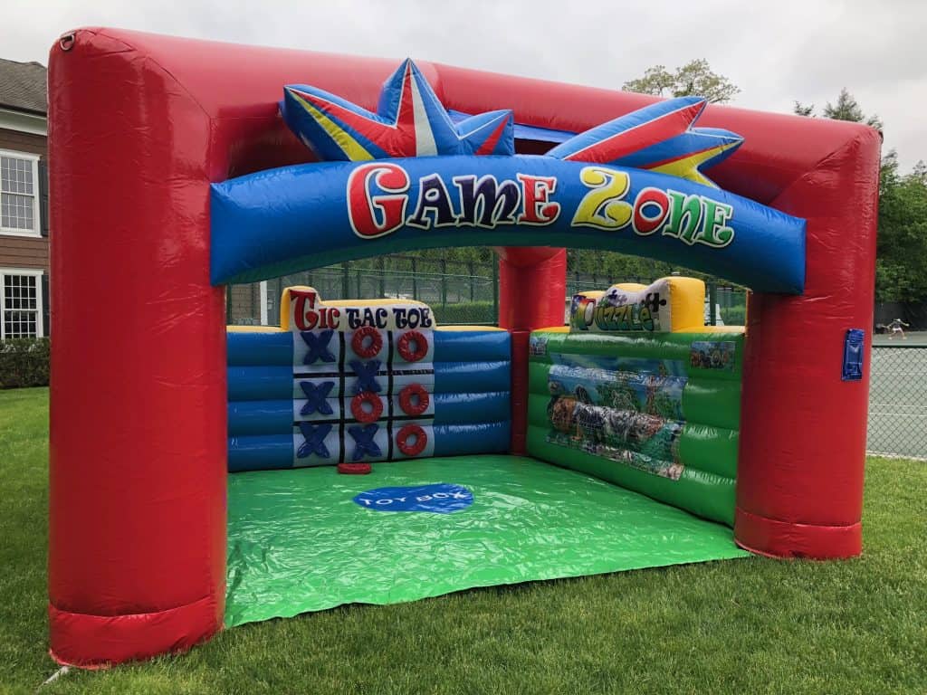An inflatable red, green, and blue game zone structure with tic-tac-toe and other activities inside is set up on a lawn near a tennis court and brick building—perfect for party rentals Long Island, NY!.