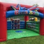 An inflatable red, green, and blue game zone structure with tic-tac-toe and other activities inside is set up on a lawn near a tennis court and brick building—perfect for party rentals Long Island, NY!.