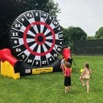 Three children in swimsuits stand on grass, aiming soccer balls at a large inflatable dartboard outdoors—perfect for party rentals Long Island, NY. Black, white, and red rings with numbers are visible, along with trees and a tennis court in the background.
