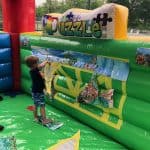 A young boy plays with a large inflatable puzzle featuring animal images, attaching pieces to a green and yellow wall inside an outdoor bounce house Long Island. Trees and a fence are visible in the background at this NY party rental event.