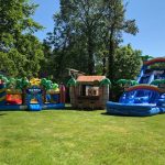 Three large inflatable attractions—a bounce house Long Island, a tiki hut, and a blue water slide with palm tree decorations—are set up on a sunny, grassy lawn surrounded by trees, perfect for memorable party rentals Long Island, NY.