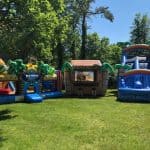 Three colorful inflatable attractions—a beach maze, tiki stand, and water slide—are set up on a sunny green lawn, surrounded by trees. Perfect for party rentals Long Island, NY, these attractions promise endless fun for all ages.