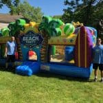 Two people stand by a colorful "Beach Party" bounce house Long Island setup, decorated with palm trees and beach balls, on a sunny lawn surrounded by trees and houses—perfect for party rentals Long Island, NY.