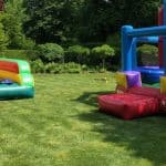 Two colorful inflatable bounce houses sit on a green lawn surrounded by trees and bushes—perfect for party rentals Long Island, NY. One bounce house features slides, while the other offers an enclosed play area for endless fun.