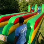 Three people enjoy a colorful inflatable bungee run game outdoors; two race along parallel lanes while a staff member in a blue shirt observes. Surrounded by green trees and grass, it's perfect for party rentals Long Island, NY.