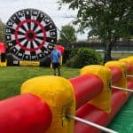 An inflatable human foosball court is set up on grass, with a large inflatable dartboard and a bounce house Long Island in the background. A person stands near the dartboard under a tree on a sunny day.