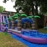 An inflatable water slide with tropical palm tree decorations set up on grass near a brick building, perfect for party rentals Long Island, with a blue tarp underneath and green trees in the background.