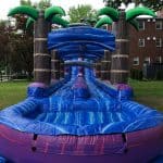 A large inflatable water slide with palm tree decorations sits on grass near a brick building in NY, featuring a blue slide lane and a small pool at the end—perfect for party rentals Long Island events.
