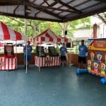 Three people in blue shirts stand by carnival game booths with red-and-white striped tents and an inflatable clown toss game, set up under a pavilion. This festive NY scene showcases party rentals Long Island style, with trees and buildings in the background.
