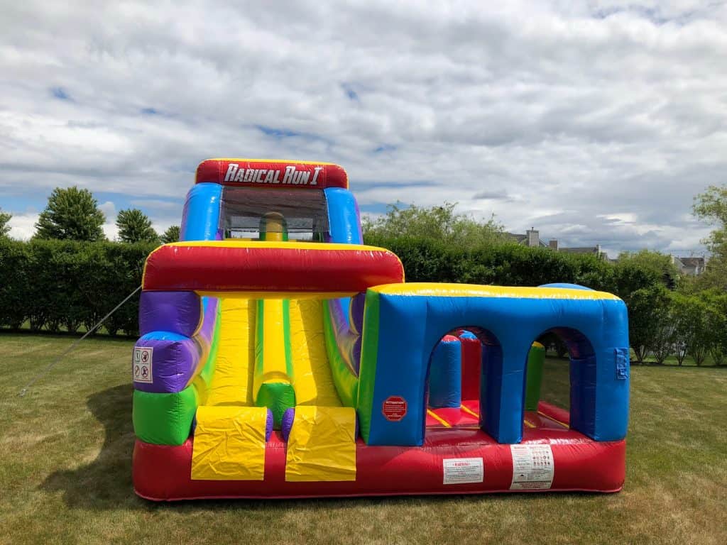A colorful inflatable obstacle course called "Radical Run" is set up on a grassy lawn, perfect for party rentals Long Island, NY. The setup features a slide, tunnels, and climbing elements with trees and bushes under a partly cloudy sky.