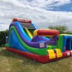 A large, colorful inflatable obstacle course and slide sits on a grassy field under a partly cloudy sky. This vibrant bounce house Long Island option features bright red, blue, yellow, green, and purple sections.