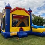 A colorful inflatable bounce house Long Island, shaped like a castle, sits on a grassy lawn in front of a house, featuring blue, yellow, and red sections with an entrance ramp. The sky is partly cloudy—perfect for NY parties!.